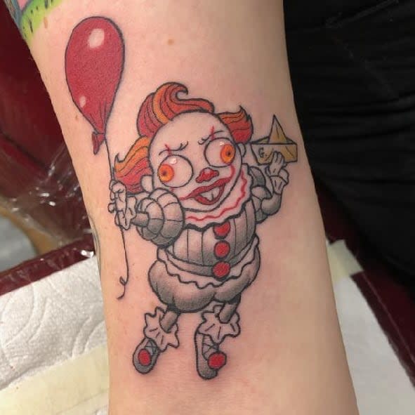 Colorful, new school Pennywise tattoo on forearm, holding a paper boat and balloon
